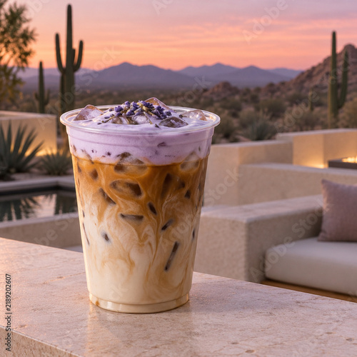 Iced lavender latte in a plastic cup with purple foam and dried flowers, set against a scenic desert sunset landscape with cacti and mountains