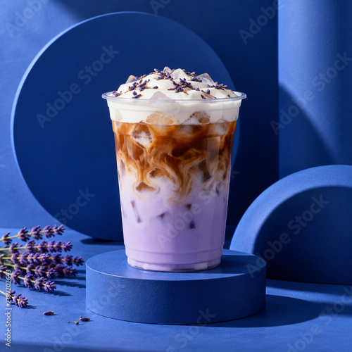 Iced lavender latte with cold foam topping in a clear plastic cup on a blue pedestal with lavender sprigs against a vibrant blue studio background