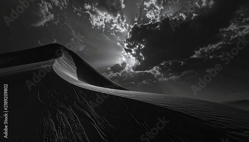Dramatic grayscale vast sand dune stretches under a dynamic, cloudy sky, illuminated by radiant sunbeams