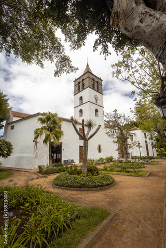 The historic Church of San Marcos, a white-washed building with a prominent bell tower and clock, situated in a green garden square in Icod de los Vinos, Tenerife, Spain.