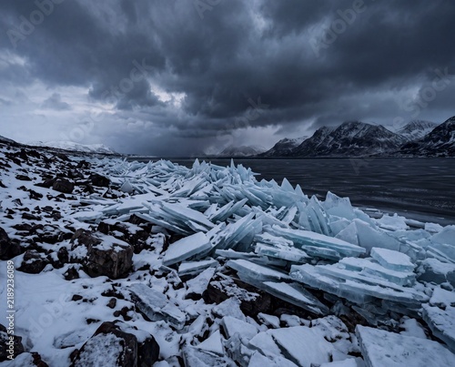 Sharp broken ice fragments piled along a frozen rocky shoreline beneath dark moody storm cloud skies