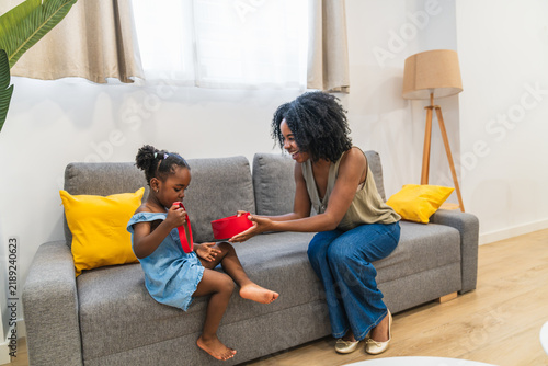 фотографія African american mother smiling, giving a gift box to her daughter sitting on a