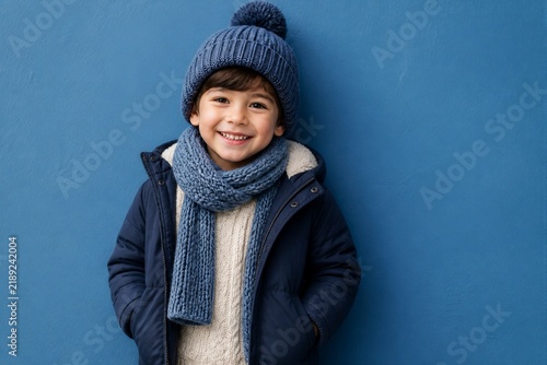 Young boy smiling in winter clothes against blue wall