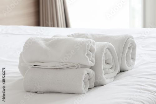 Rolled clean white cotton towels neatly stacked on a soft white bed sheet in a hotel room, symbolizing hospitality, spa relaxation, and luxury comfort.