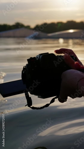 A videographer holds a mirrorless camera with an open screen above water during golden hour. Essential visual asset for social media vlogging, content creation, and digital filmmaking.