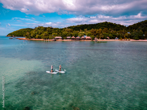 View of couple of tourist are enjoy on water sport paddle sup-board with kayaking along at the edge of the sea beach under crystal clear of water in the island of summer trip