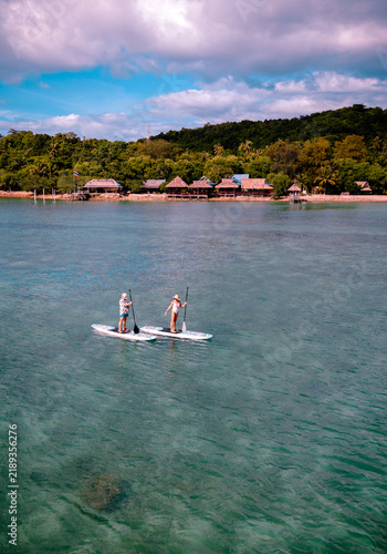 View of couple of tourist are enjoy on water sport paddle sup-board with kayaking along at the edge of the sea beach under crystal clear of water in the island of summer trip