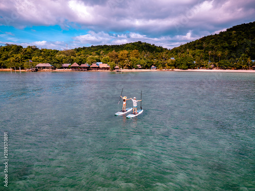 View of couple of tourist are enjoy on water sport paddle sup-board with kayaking along at the edge of the sea beach under crystal clear of water in the island of summer trip