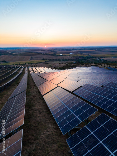 Solar panels in a large field capture sunlight at sunset on the horizon with rolling hills in the background