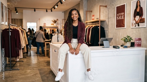 Smiling Black female fashion boutique owner sitting at checkout counter during a seasonal clothing sale