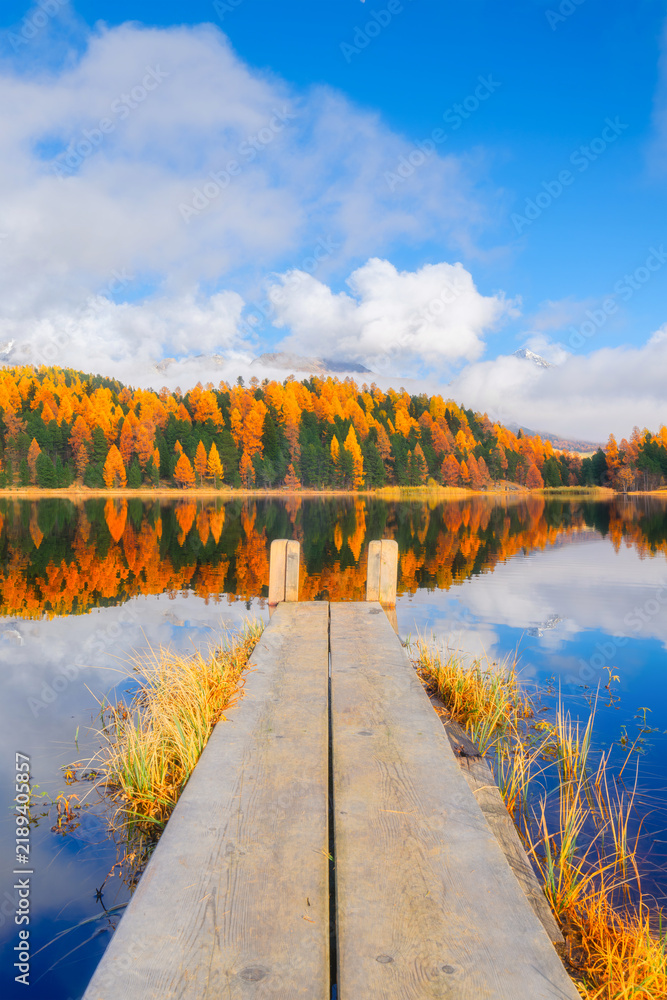 Fototapeta premium Vertical photo. Lake and a small pier. Autumn landscape. Clouds and fall forest. Incredible view of the mountain valley with lake. Background, wallpaper, postcards