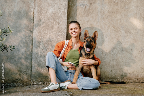 Фотография Happy young woman hugging her german shepherd dog while sitting on the street near a concrete wall