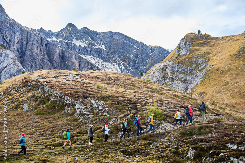 Obraz premium Group of hikers climbs rocky alpine trail across open Dolomites terrain, surrounded by steep ridges, autumn grass, scattered snow. Conveys teamwork, endurance, active travel, guided outdoor adventure