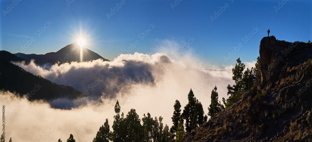 Obraz premium Silhouetted man photographs Mount Teide sunset with smartphone from cliff above panoramic sea of clouds in Tenerife, Spain. Travel photography scene combines adventure, exploration, volcanic scenery