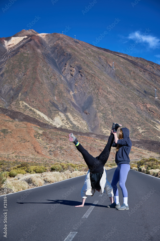Fototapeta premium Female traveler helps friend hold playful handstand on mountain road near Mount Teide, expressing trust, teamwork and active friendship during Tenerife adventure in Canary Islands, Spain