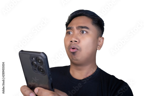 Obraz na plátne Surprised and startled Indonesian man in a black t-shirt looking at his smartphone with a rounded open mouth and wide eyes against a white background