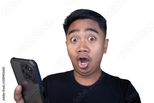 Obraz na plátne Surprised and stunned Indonesian man in a black t-shirt looking at his smartphone with wide eyes and an open rounded mouth against a white background