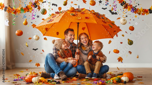 Happy family of four sitting together indoors under orange umbrella while autumn leaves and pumpkins fall