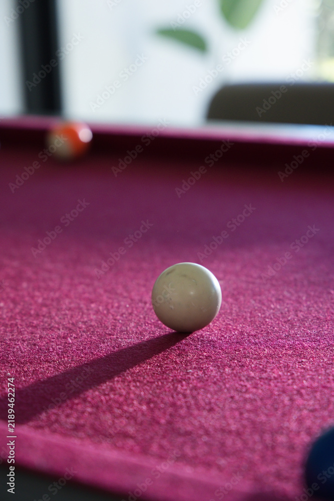 Fototapeta premium White cue ball is resting on red felt pool table, casting long shadow toward lower-left
