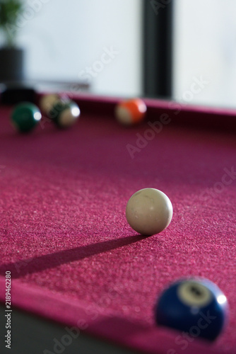 White cue ball sitting sharp on burgundy felt pool table near vertical window, blurred balls