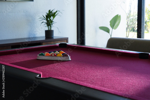 Magenta felt pool table holding metal triangle rack with colorful balls near windowed sofa