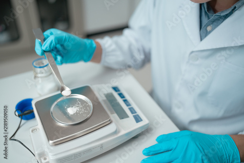 Scientist weighing white powder sample on precision scale using tweezers in laboratory setting