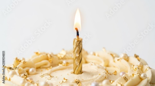 Golden Birthday Candle Burning on White Frosted Cake with Gold Flakes and Pearls, Close-up