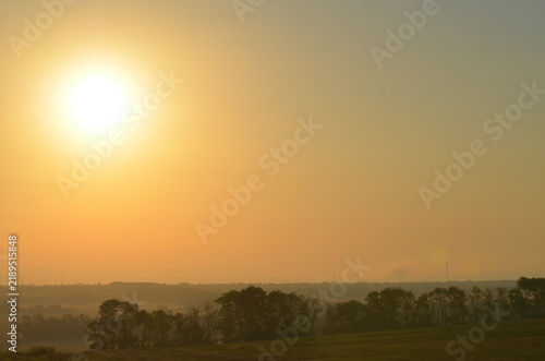 Fotografie A beautiful scenic view of a bright hazy sun rising over rolling agricultural fields and green trees at sunrise
