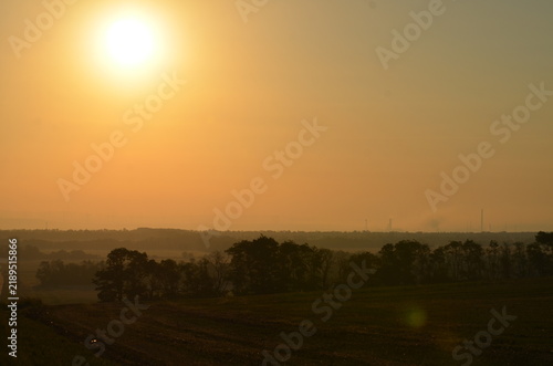 Obraz na płótnie A beautiful scenic view of a bright hazy sun rising over rolling agricultural fields and green trees at sunrise