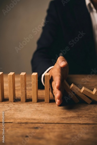 Business person hand stopping falling wooden domino blocks on table for risk management concept