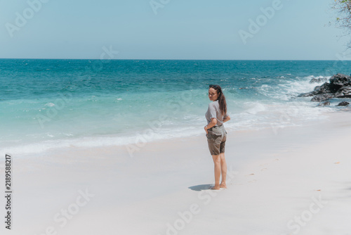 Obraz na płótnie Full length portrait of an Asian woman standing on white sandy beach near ocean waves looking over shoulder towards camera