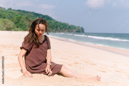 Obraz na płótnie Asian woman in brown dress sitting on white sandy beach playing with sand while looking down under warm bright daylight
