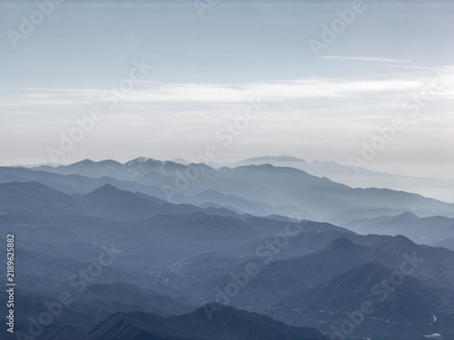 Aerial view of layered blue-gray mountain ridges fading into haze, distant peaks dusted with snow under a soft sky near Santa Clarita, California