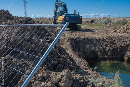 Quadro su tela A metal safety fence borders an excavated earthen pit filled with water, where a tracked excavator stands working on a sunny construction site