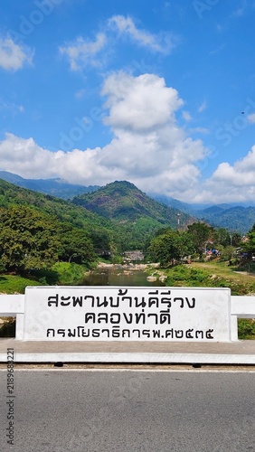 Картината върху платно A white bridge sign overlooking the Tha Di Canal, framed by lush green mountains, a quiet river, and dramatic white clouds beneath a vivid blue sky in Khiriwong, Thailand