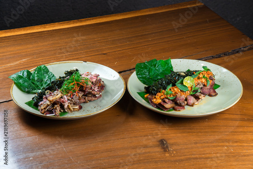 Gourmet Delicacy on Plates: Two exquisite dishes featuring thinly sliced meat, garnished with fresh green leaves, chili, and herbs, presented on ceramic plates atop a rustic wooden table.