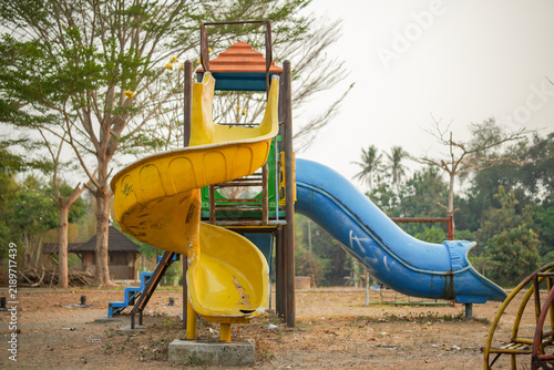 An abandoned playground featuring a worn yellow spiral slide and a blue straight slide set in a dry park among trees and faded equipment