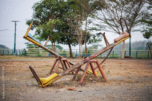 A rusty abandoned seesaw with yellow seats sits on an empty outdoor playground surrounded by dry grass and trees, suggesting nostalgia, neglect and quiet urban decay.
