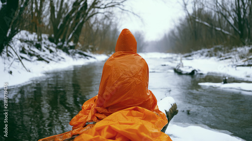 Person in emergency blanket sitting on snowy log by frozen stream, hypothermia and winter survival concept