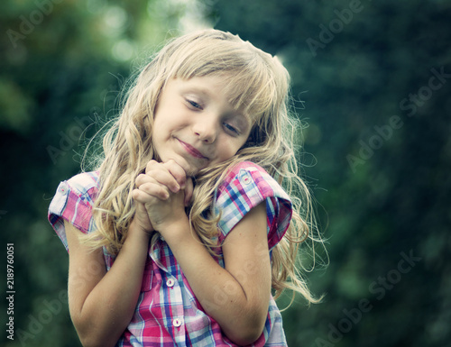 Foto Cute little blonde girl with wavy hair clasping her hands together with a sweet gentle smile outdoor against a natural green foliage background