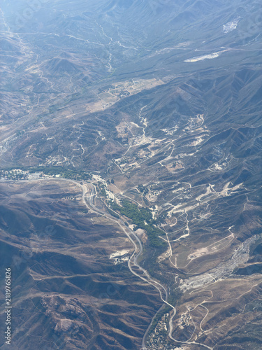 Aerial shot of a dry mountain valley with winding roads, scattered structures, and sun-bleached washes cutting through hillsides, Agua Dulce area, California