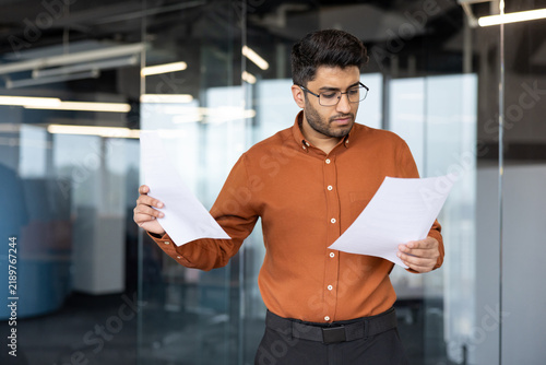 Картината върху платно Young businessman in orange shirt studies printed documents inside modern office