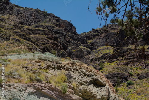 Landscape perspective showing the distinctive geological strata, sparse brush, and towering mountain slopes under bright daylight.