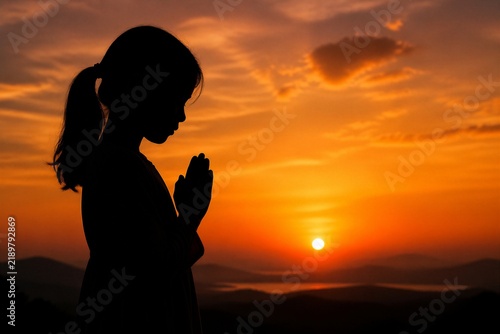 Silhouette of a person praying at sunrise. Hands clasped in quiet devotion. Golden light paints the sky with warm hues. Distant hills fade into the horizon's glow