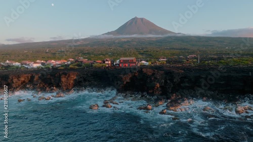 Aerial view of a coastal village on Pico Island, Azores, featuring traditional houses on rugged volcanic cliffs, with the iconic Mount Pico volcano and a visible moon in the clear sky.