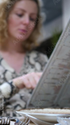 Woman reads menu at restaurant table, studying choices with focused calm. Her hand points along printed text while soft light and shallow focus create quiet dining atmosphere during meal ordering.