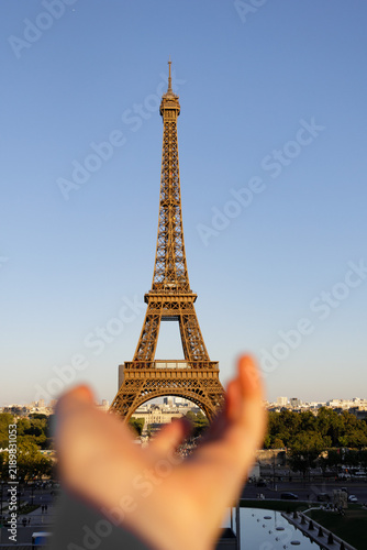 Outstretched hand reaches toward the Eiffel Tower during golden hour in Paris, France, creating a playful travel concept with the iconic landmark in clear focus.