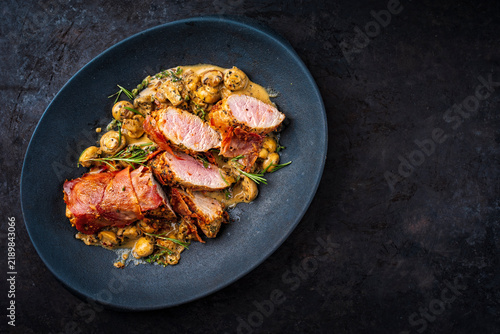 Pork fillet roast wrapped in prosciutto ham bacon with mushroom cream sauce and fresh herbs served as top view in a cast iron skillet