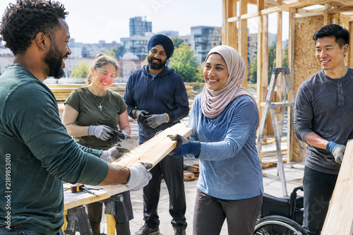 Diverse community volunteers building house together, smiling multicultural group carrying wood planks, inclusive teamwork at construction site