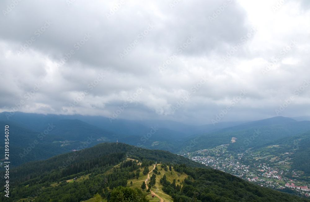 Fototapeta premium Expansive mountain landscape with rolling green hills, a winding ridge trail and a small village nestled in the valley beneath dramatic low clouds and moody overcast skies. Carpathians, Ukraine